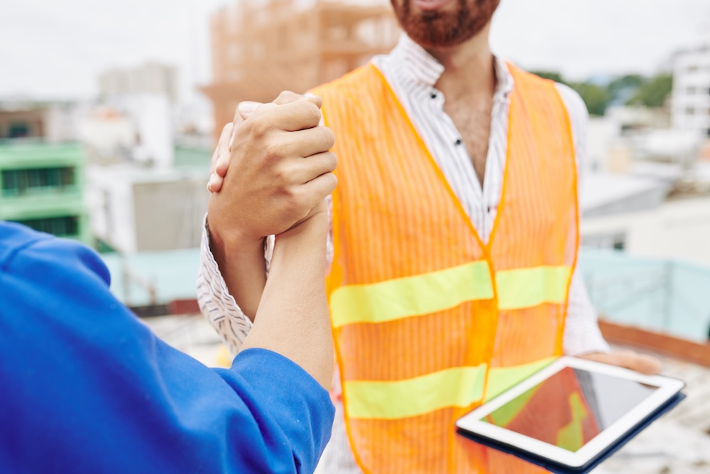 A construction worker in a safety vest shakes hands with someone on a building site, with one holding a digital tablet.