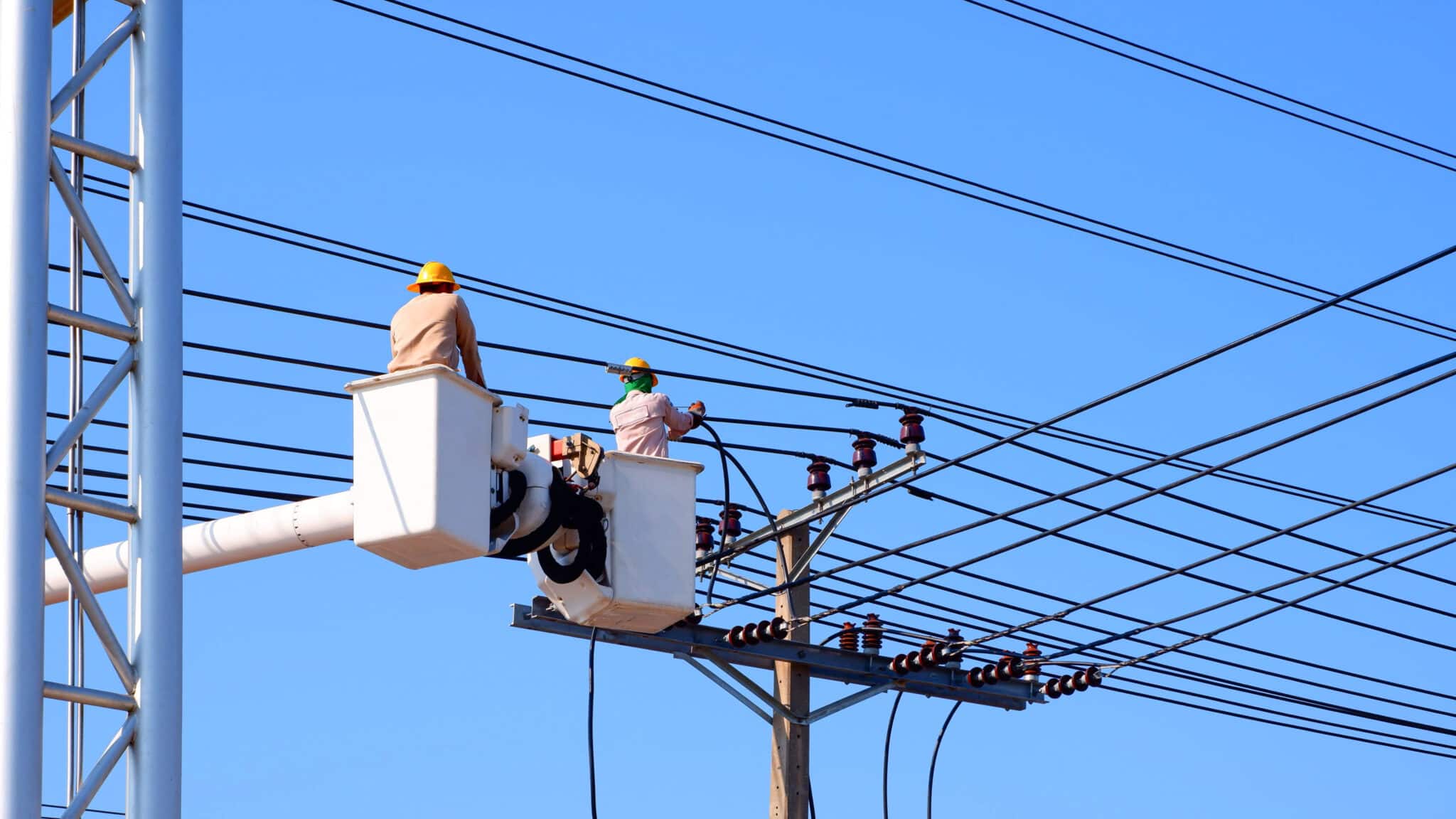 Two electricians wearing safety helmets and vests working on power lines from a bucket boom truck.