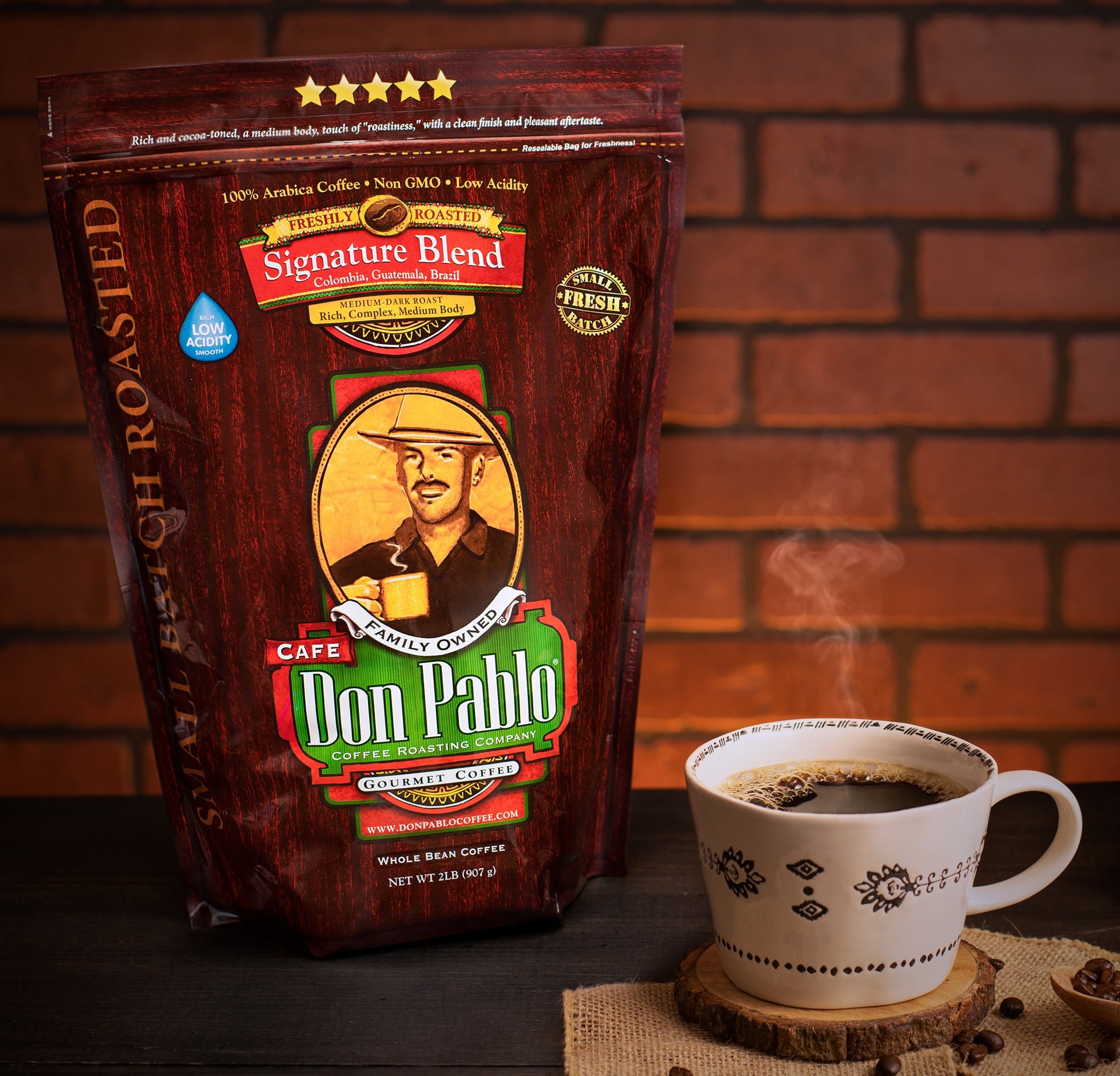 A bag of Don Pablo Signature Blend coffee beans next to a steaming mug of coffee, with a brick wall in the background.