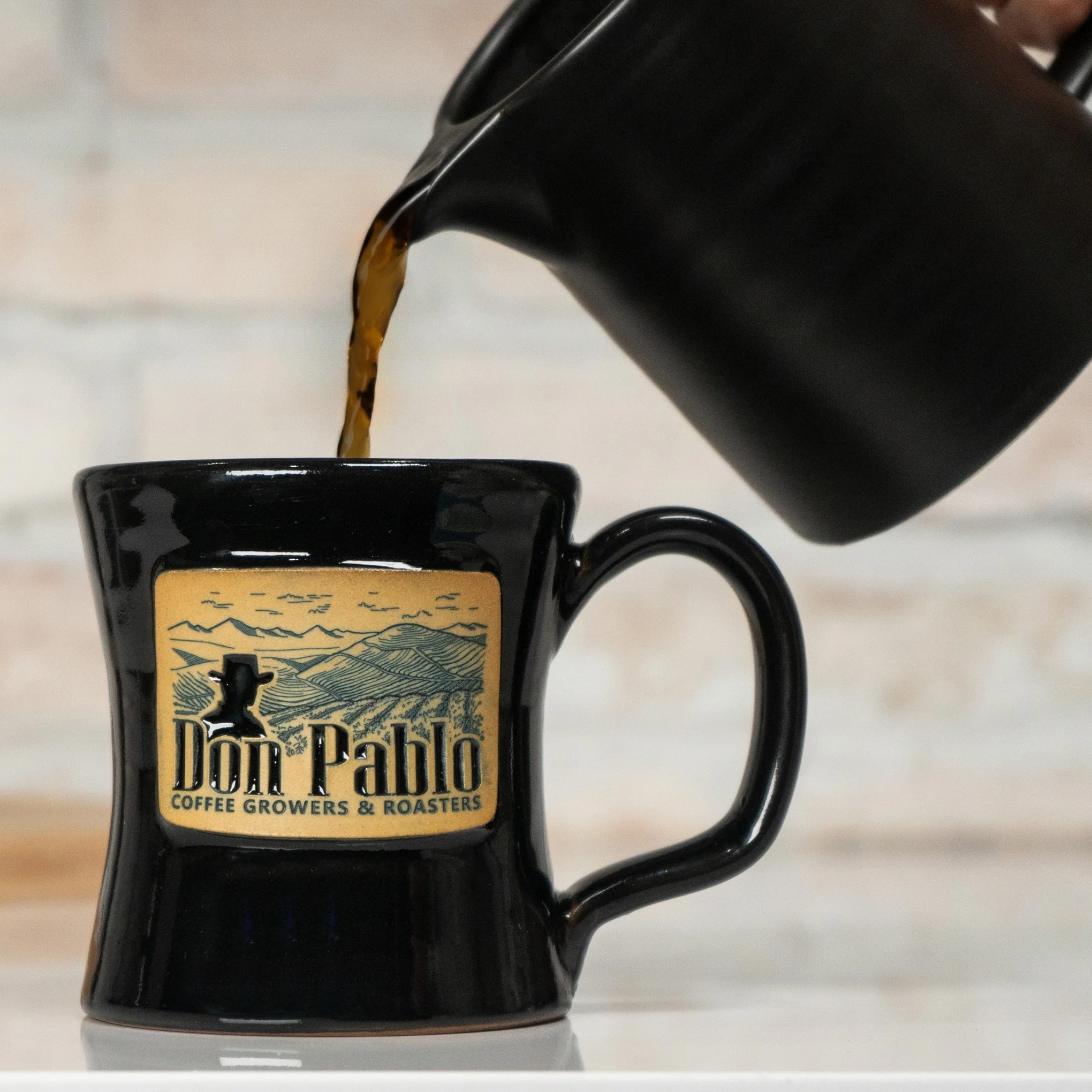 Coffee being poured into a black mug with "Don Pablo Coffee Growers & Roasters" logo on it, against a brick wall backdrop.